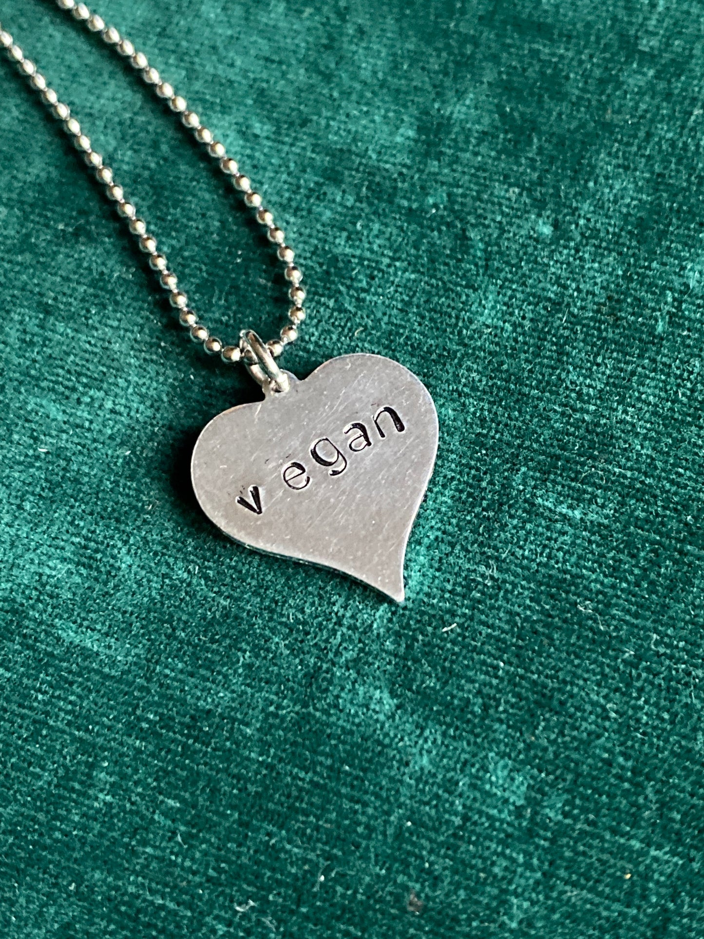 This is a proximately 1 inch heart-shaped aluminum pendant hung on a ball drain chain, and it says vegan in lowercase letters stamped onto the pendant